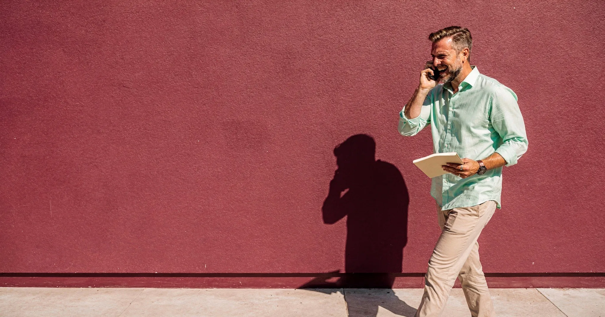 Un account manager rit au téléphone en marchant devant un mur framboise au soleil